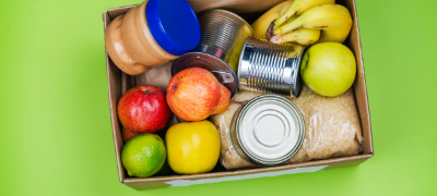 Cardboard box overhead shot showing canned food, fruits and peanut butter jar.