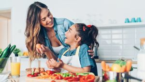 A smiling woman and a young girl in an apron laugh together in a bright kitchen while preparing food with colorful fresh vegetables and eggs.