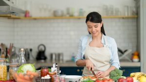 A woman wearing an apron is carefully dicing vegetables on a cutting board, demonstrating food safety practices.
